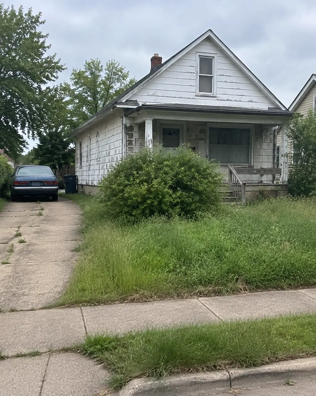 A worn older house with peeling paint, an overgrown yard, and an older car in the driveway